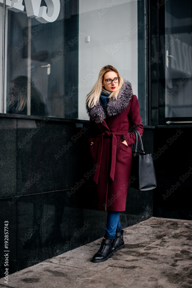 Young girl with blonde hair is standing in a burgundy coat and fur collar near the storefront. Fashion shooting Young girl with blonde hair is standing in a burgundy coat and fur collar near the storefront. Fashion shooting