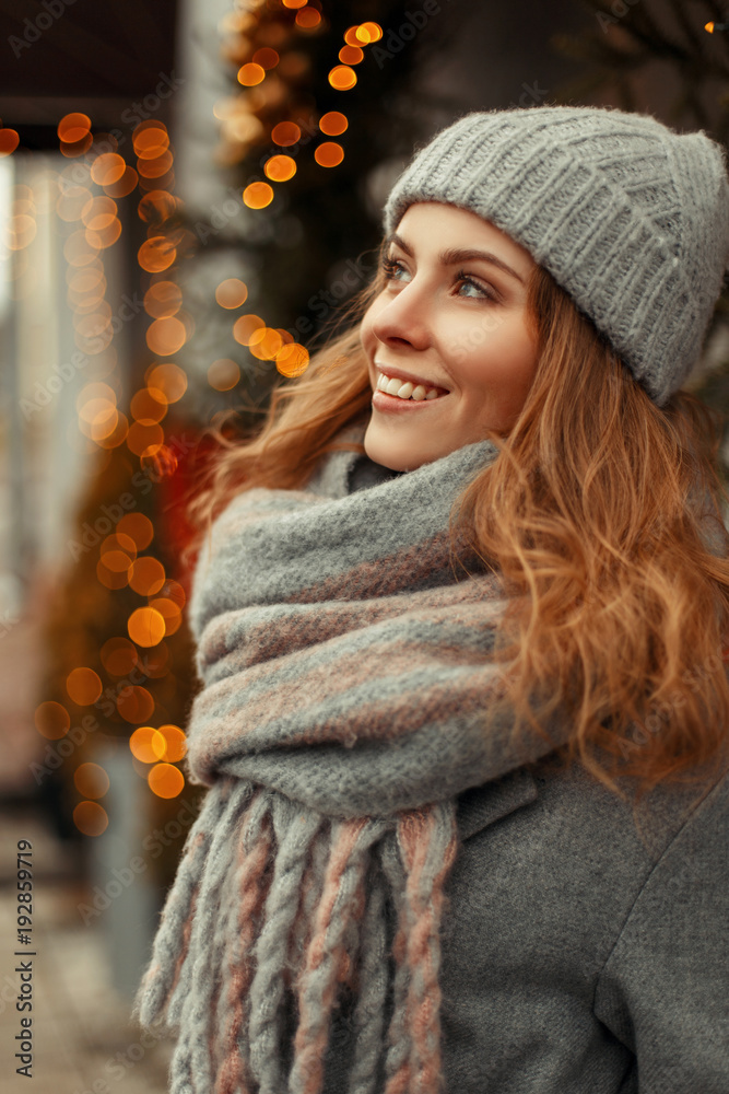 Happy young woman with a magic smile in fashion stylish knitwear clothes  with a knitted gray hat and scarf near the lights on a holidays фотография  Stock | Adobe Stock