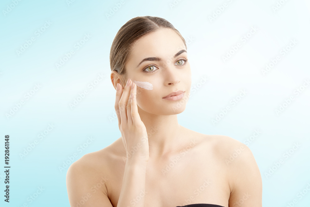 Studio shot of beautiful young woman applying moisturizer cream onto her face against at isolated light blue background with copy space. 
