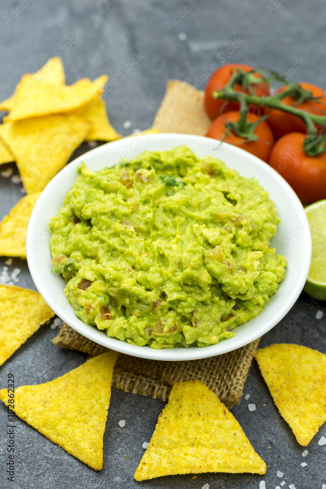 Avocado dip guacamole with tortilla chips in a white bowl on a black stone table. Traditional Mexican appetizer.