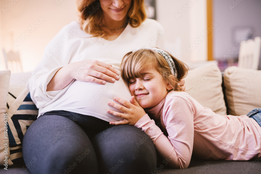 Cute smiling little toddler girl embracing belly of her pregnant mother while sitting on the sofa. Family and new life concept. Cute smiling little toddler girl embracing belly of her pregnant mother while sitting on the sofa. Family and new life concept.