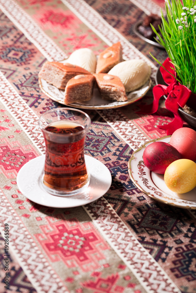 Fototapeta premium Novruz setting table decoration, tea in tulip shape glass on ethnic motives rustic table cloth with wheat grass, dyed eggs, traditional sweets, samovar, new year sring celebration, nature awakening