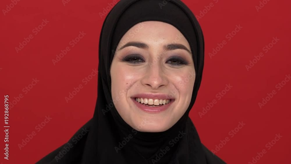 Portrait of middle eastern woman with smoky eyes visage putting off black niqab or burqa and smiling on camera, over red background. Concept of emotions Portrait of middle eastern woman with smoky eyes visage putting off black niqab or burqa and smiling on camera, over red background. Concept of emotions
