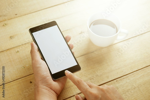 Hand holding black mobile smart phone with blank screen with coffee cup on wood table . technology and lifestyle concept.