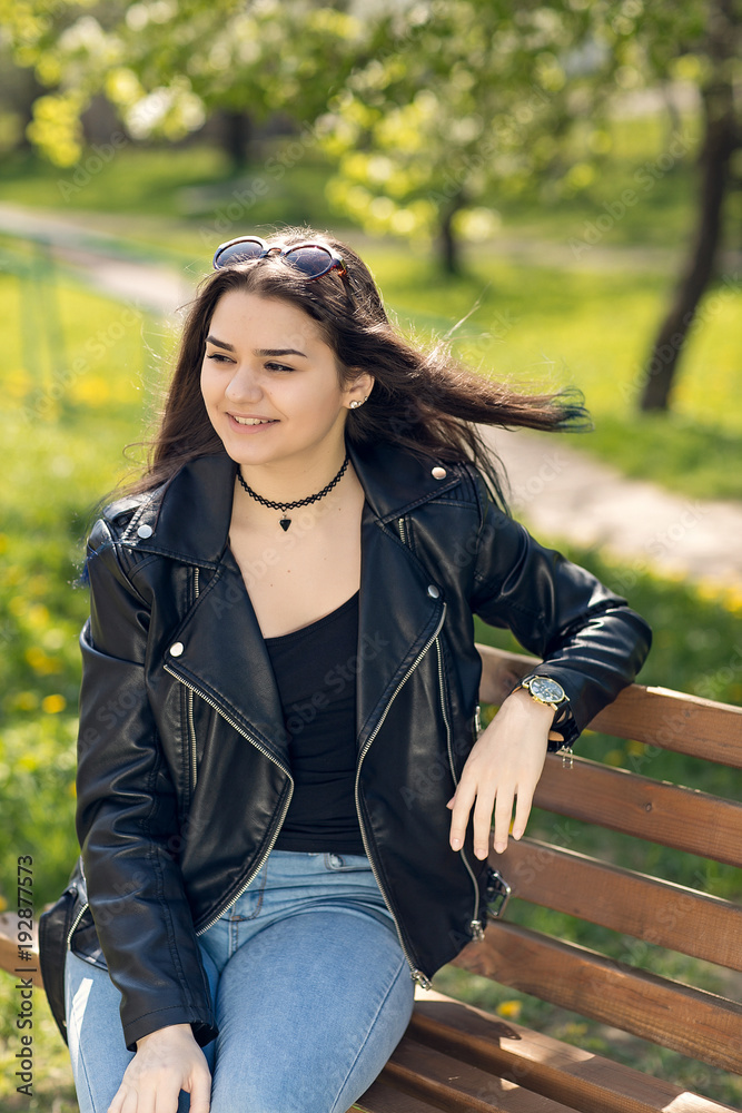 Glamorous young Caucasian woman in black leather jacket sitting in the park on the bench Glamorous young Caucasian woman in black leather jacket sitting in the park on the bench