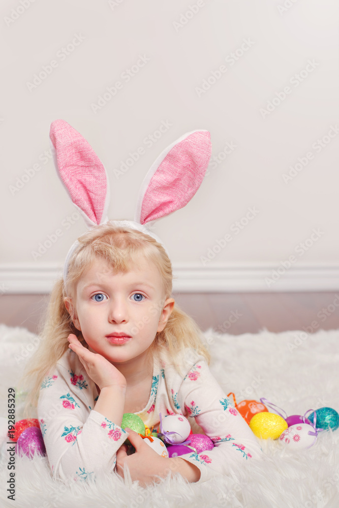 Cute adorable Caucasian child girl wearing Easter bunny rabbit ears lying on soft fluffy rug carpet in studio. Kid holding holiday colorful eggs celebrating traditional holy Christian holiday Cute adorable Caucasian child girl wearing Easter bunny rabbit ears lying on soft fluffy rug carpet in studio. Kid holding holiday colorful eggs celebrating traditional holy Christian holiday