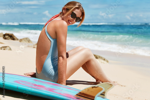 Adorable beautiful young female model wears sunglasses and bathing suit, sits on hot sandy beach against ocean background, sunbathes and smiles happily, enjoys good vacation in tropical country © wayhome.studio 
