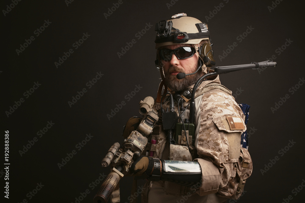 Brutal man in the military desert uniform and body armor stands in a fighting rack and holds his rifle on a black background. The bearded player in the airsoft safety glasses aiming a rifle