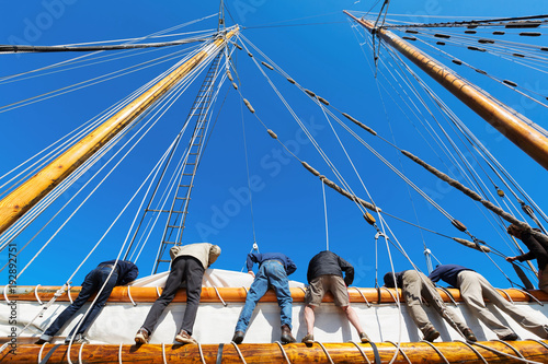 Crew leans over the side of a big sailboat to raise the heavy sail on a tall ship at sea. Takes six strong people to hoist the canvas sail. Theme for teamwork, cooperation