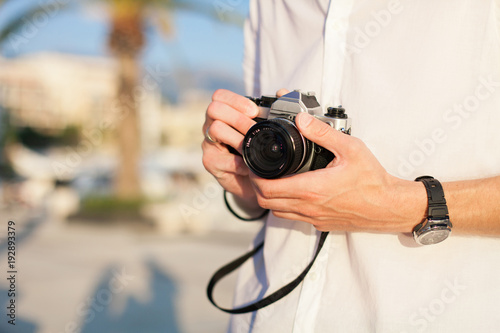 Travel photographer has film photo camera in his hand outdoor. Tourist has vacation in sea resort. Man is wearing in casual white shirt and wristwatch. Blurred background with palm and space for text.