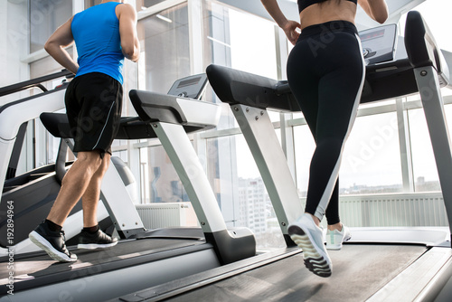 Mid-section back view of two fit young people, man and woman, running on treadmills facing windows in modern gym, copy space