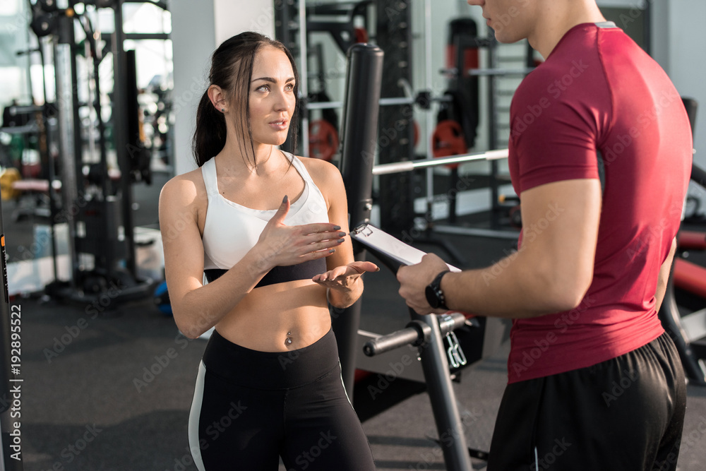 Portrait of young woman talking to personal fitness coach in modern gym, copy space Portrait of young woman talking to personal fitness coach in modern gym, copy space