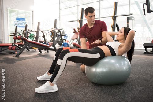 Side view portrait of muscular coach helping young woman doing exercises on fitness ball in modern gym, copy space
