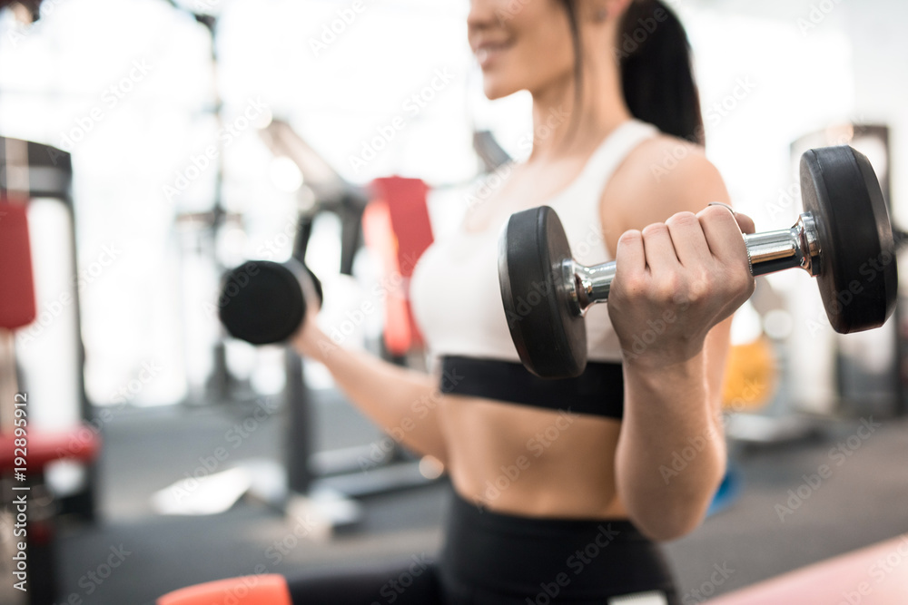 Close up of sportive young woman training with dumbbells in modern gym, focus on heavy dumbbell in hand, copy space Close up of sportive young woman training with dumbbells in modern gym, focus on heavy dumbbell in hand, copy space