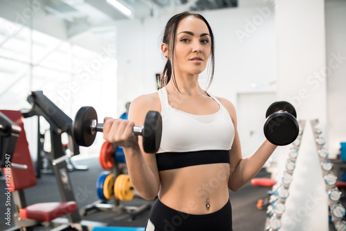 Portrait of sportive young woman training with dumbbells in modern gym and looking at camera, copy space