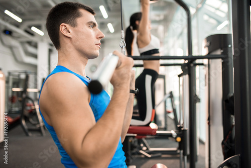 Side view of two people exercising on machines in modern gym by window, focus on handsome muscular man