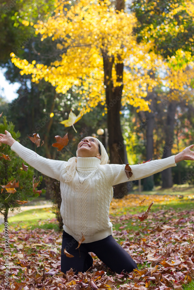 Happy mature woman on white turtle neck sweater and shawl covering her head enjoying fall season at park with open arms, knees on the ground of brown leaves and green and yellow trees in the forest