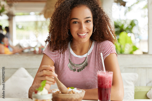 Candid shot of happy dark skinned young female with Afro hairstyle, eats delicious exotic salad and drinks smoothie, being in good mood, enjoys good rest at cafeteria. People and leisure concept