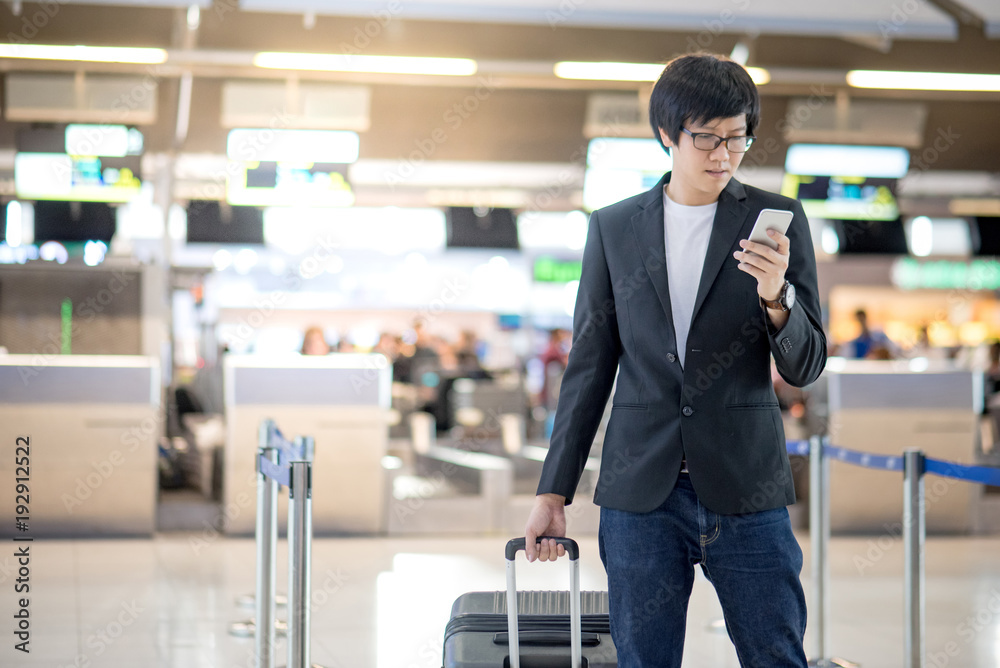 Young asian man with his suitcase luggage using smartphone while waiting for airline flight in the international airport terminal, business travel and online check in concepts