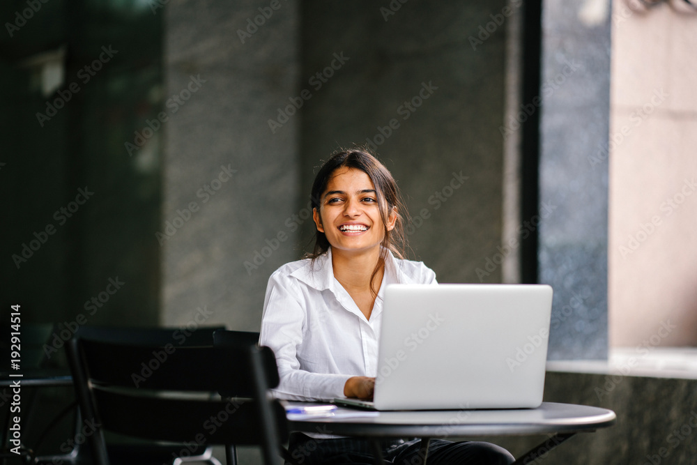 An image of a young Asian Indian woman who types and works in a laptop and seeks a job. She wears a professional, crisp white shirt and she smiles during the day as she sits at a table. An image of a young Asian Indian woman who types and works in a laptop and seeks a job. She wears a professional, crisp white shirt and she smiles during the day as she sits at a table.