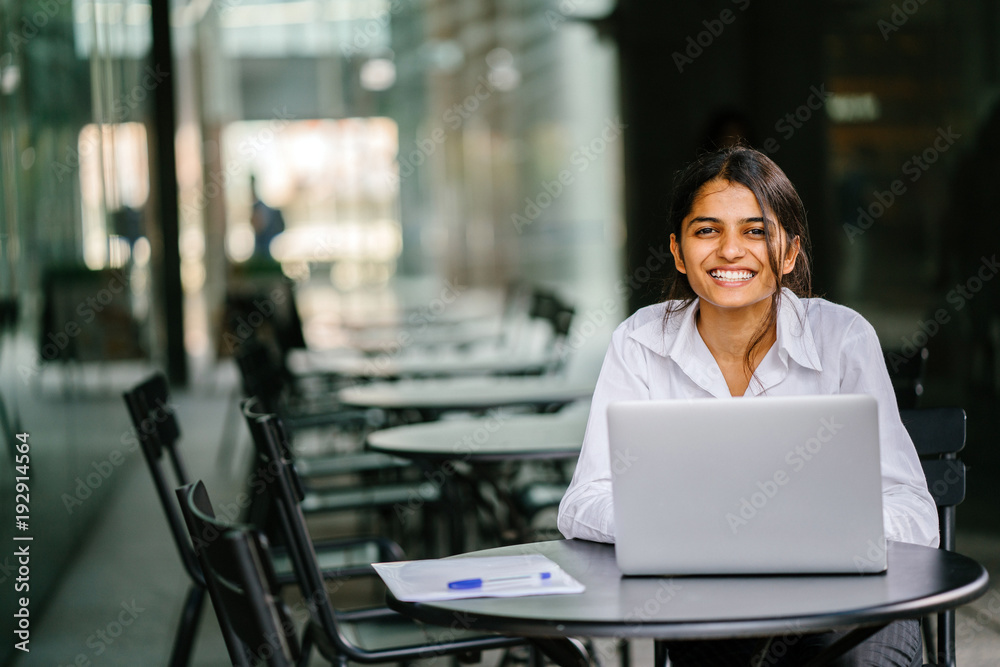 A young Indian Asian woman is typing and working in a laptop and looking for a job during the day She is wearing a professional, crisp white shirt and she is smiling as she types on her laptop A young Indian Asian woman is typing and working in a laptop and looking for a job during the day She is wearing a professional, crisp white shirt and she is smiling as she types on her laptop