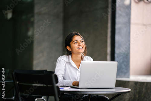 A portrait of a young Asian Indian woman sitting at a table in search of a job on her laptop. She smiles as she thinks and seems to enjoy looking for her future job.