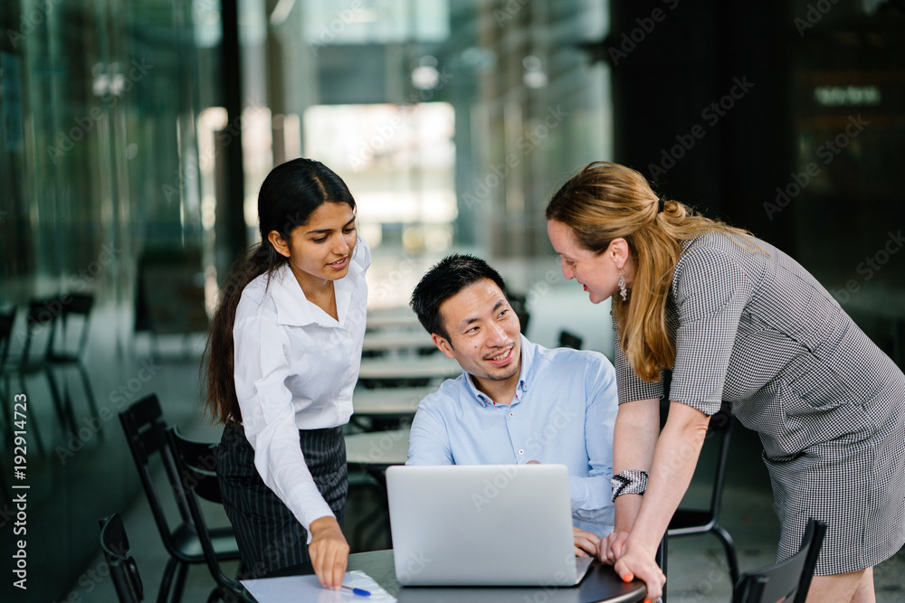 A diverse multi-racial team of three colleagues businesspeople have a relaxed discussion around a laptop notebook computer in an office or coworking space. They are all smiling and talking animatedly. A diverse multi-racial team of three colleagues businesspeople have a relaxed discussion around a laptop notebook computer in an office or coworking space. They are all smiling and talking animatedly.