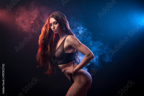 Young beatiful woman in black underwear posing against a background of blue and red smoke from a vape on a black isolated background
