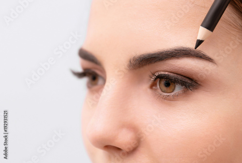 Young woman correcting shape of eyebrows on light background, closeup