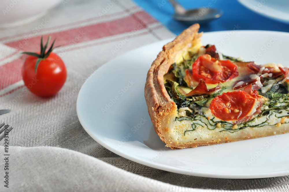 Portion of French pie quiche on a white plate. Filling with spinach, bacon, cheese, cherry tomato. In the frame, a cherry tomato. The background is draped with a napkin. Close-up. Macro photography. Portion of French pie quiche on a white plate. Filling with spinach, bacon, cheese, cherry tomato. In the frame, a cherry tomato. The background is draped with a napkin. Close-up. Macro photography.