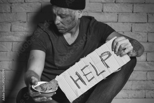 Homeless poor man holding carton board with word HELP and bowl for donations while sitting near brick wall