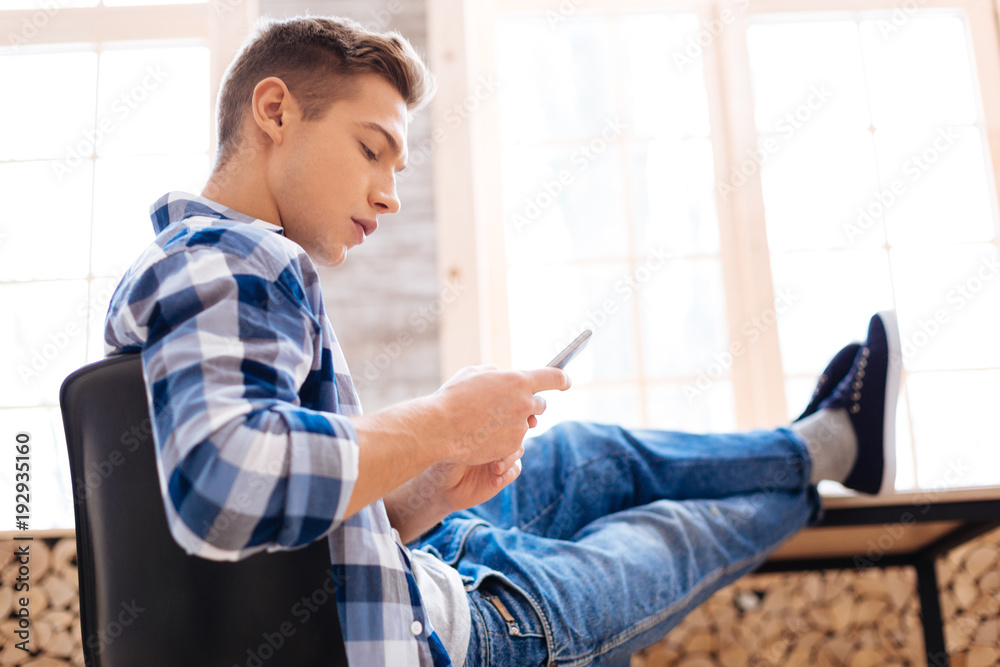 Being serious. Attractive concentrated fair-haired boy sitting with his feet on the table and using his phone and wearing a tartan shirt