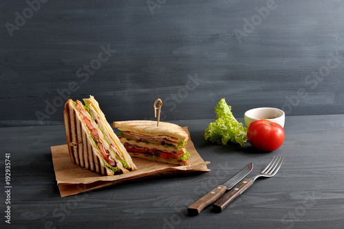 Two halves of the club sandwich on kraft paper. The filling of the sandwich consists of ham, cheese, tomato, bacon, sauce and fried egg. Nearby cutlery, mustard, vegetables. Dark background. Close-up.
