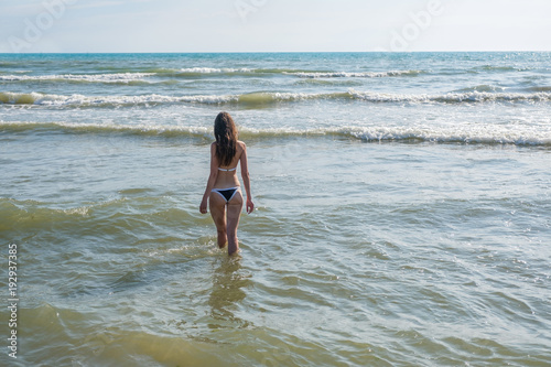 Unrecognizable woman with long hair in blue bikini walking down the water on the seaside. Back view.