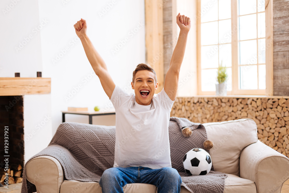 Football fan. Good-looking delighted well-built adolescent smiling and  holding his arms up while sitting on the couch and a soccer ball near him  Stock Photo | Adobe Stock