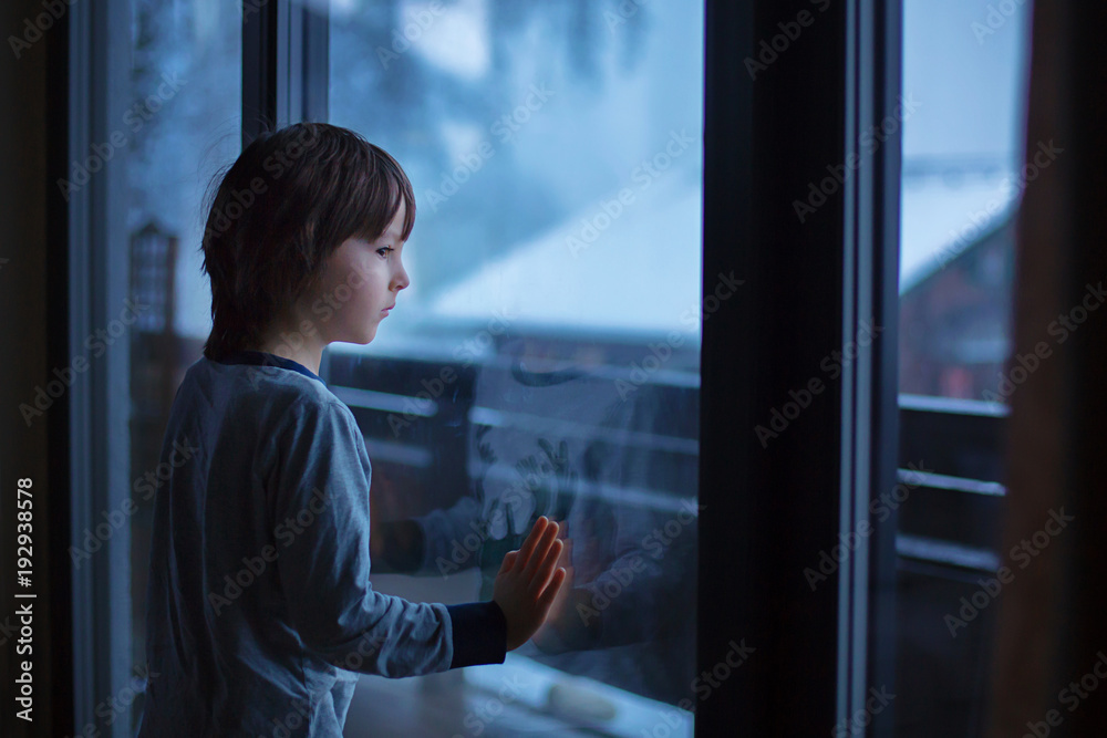 Preschool boy, sitting by the window in living room, looking at a snowy landscape
