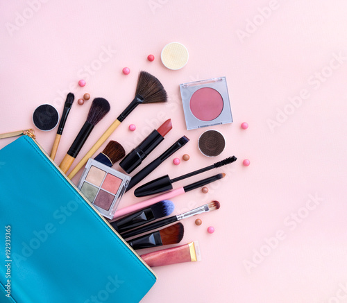 A blue cosmetics bag with makeup products spilling out on to a pastel pink background. Top view