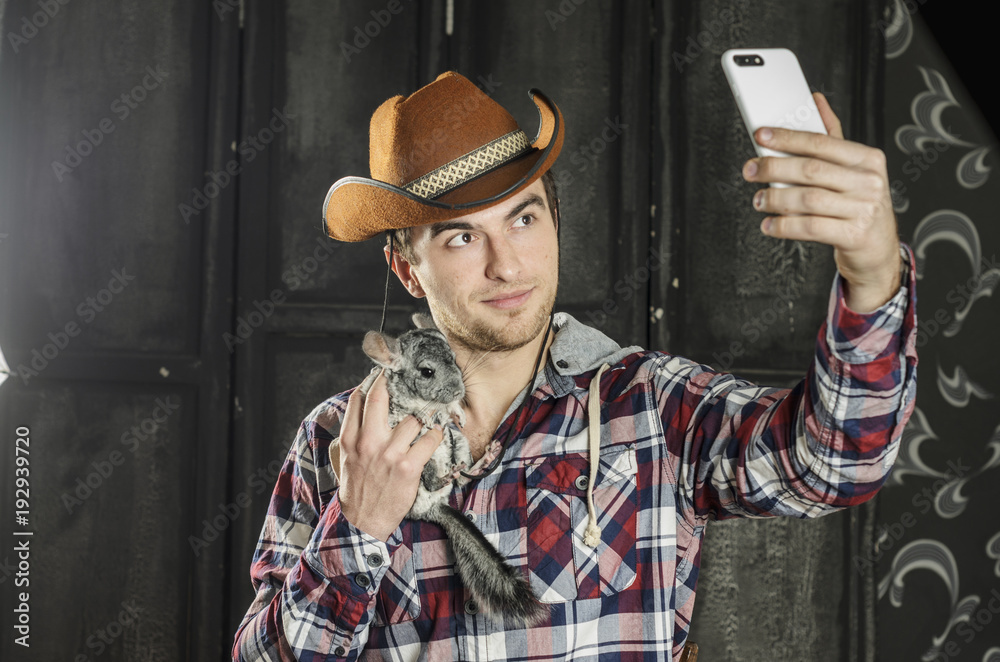 Portrait of a Cowboy. dark hair, jeans , plaid shirt. he holds the chinchilla animal . photographing self on smartphone Portrait of a Cowboy. dark hair, jeans , plaid shirt. he holds the chinchilla animal . photographing self on smartphone