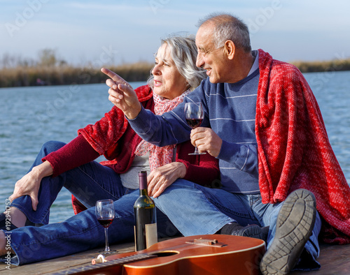 Happy senior couple enjoying time together drinking wine by the lake wrap around in a red blanket. © Zoran Zeremski