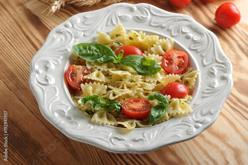 Pasta farfalle with pesto sauce and cherry tomatoes in a deep white plate. The pasta is decorated with fresh basil leaves. Light wooden background. Close-up.