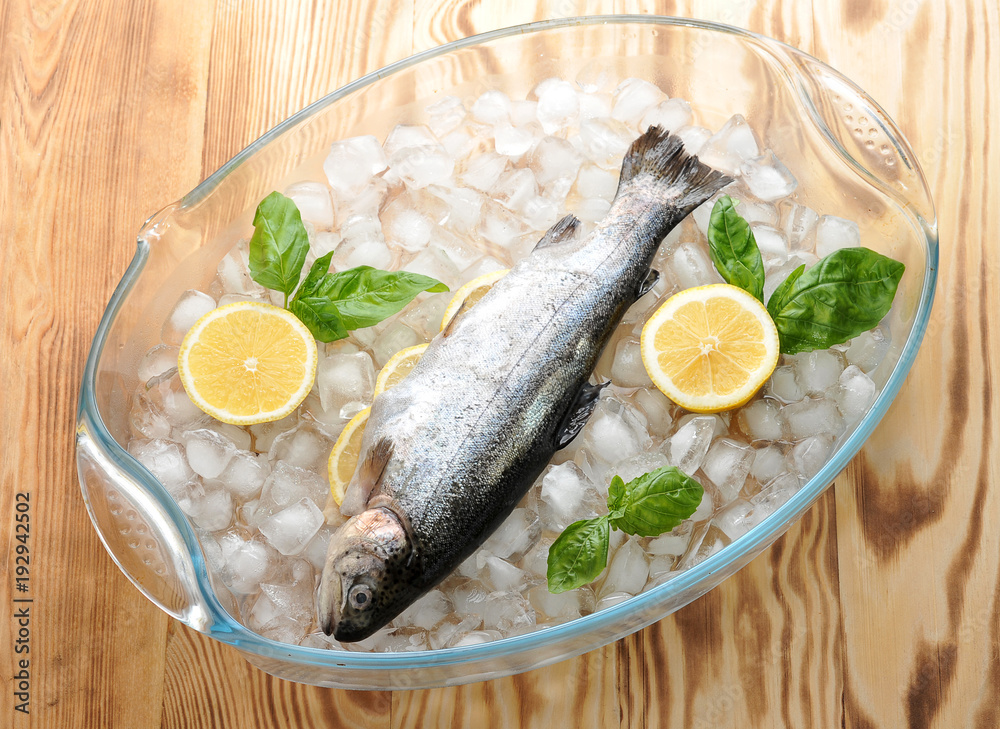 Fresh brook trout on chunks of crushed ice in a glass bowl. In the belly of trout, there are slices of lemon and rosemary. Next lemon and fresh basil. View from above. Light wooden background. Fresh brook trout on chunks of crushed ice in a glass bowl. In the belly of trout, there are slices of lemon and rosemary. Next lemon and fresh basil. View from above. Light wooden background.