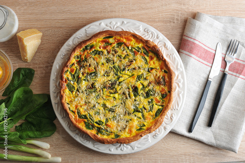 Cake pie of quiche with spinach and green onions on a white plate. Next Parmesan cheese, green onions, spinach, bowl with raw eggs without shell. Napkin made of cloth and cutlery. View from above.