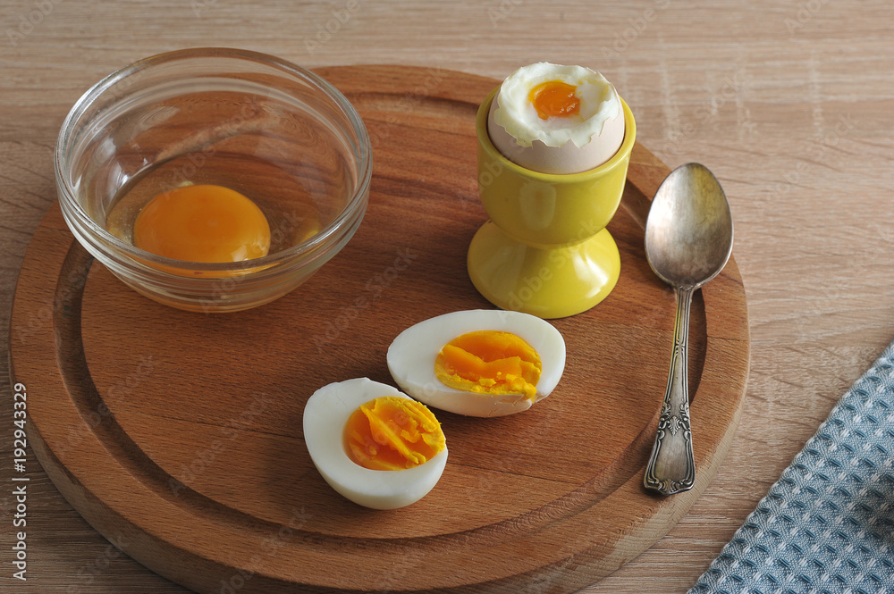 On a wooden tray are two halves of an hard-boiled eggs, an soft-boiled egg and a glass bowl with broken raw egg. Next teaspoon. Wooden light background. Closeup. On a wooden tray are two halves of an hard-boiled eggs, an soft-boiled egg and a glass bowl with broken raw egg. Next teaspoon. Wooden light background. Closeup.