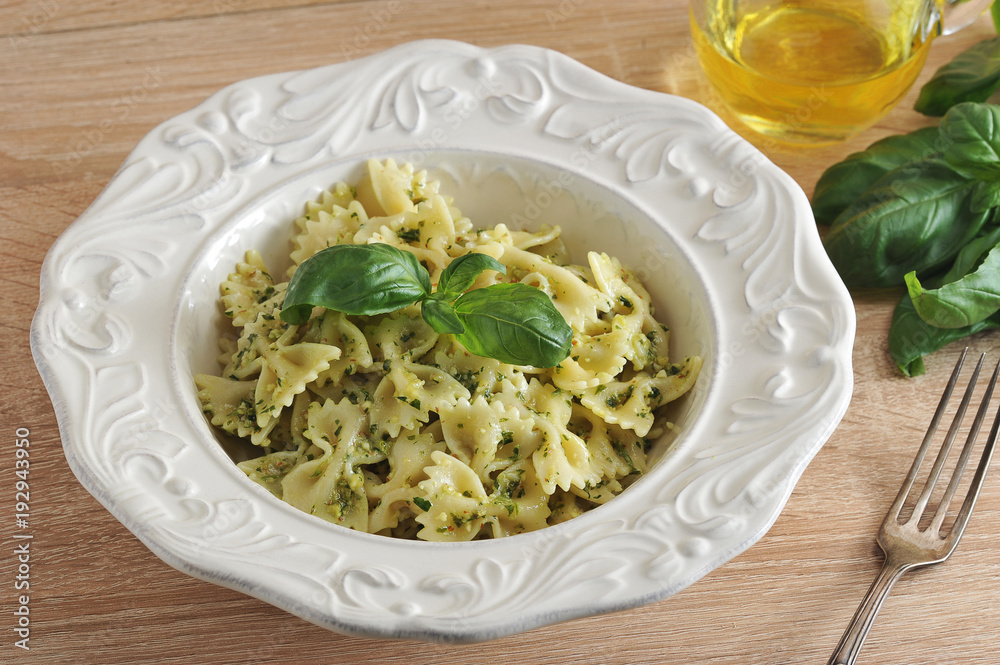 Pasta farfalle with pesto sauce in a white plate. The dish is decorated with a leaf of fresh basil. Next to the plate is a fork, a jug of olive oil and a bunch of fresh basil. Close-up.