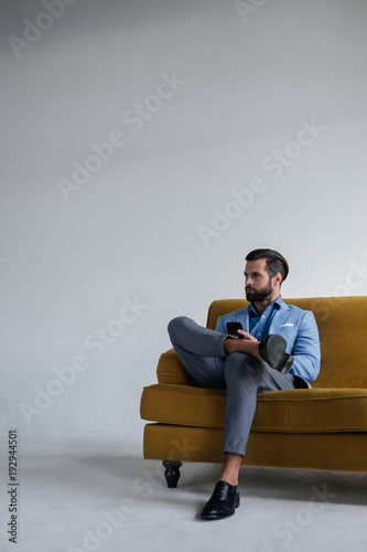 stylish man in blue trendy suit holding smartphone and sitting on yellow sofa