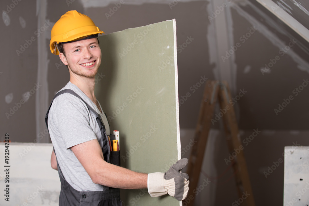 Mann auf einer Baustelle trägt eine Gipskartonplatte - obrazy, fototapety, plakaty 