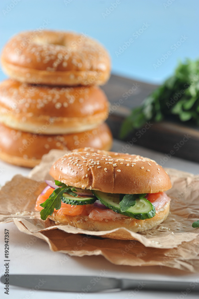 Beagle with a filling of salted salmon, arugula, cucumber, soft cheese. Beigle on kraft paper. In the background there is one more Beigle. Light background. Close-up. Macro photography. Beagle with a filling of salted salmon, arugula, cucumber, soft cheese. Beigle on kraft paper. In the background there is one more Beigle. Light background. Close-up. Macro photography.