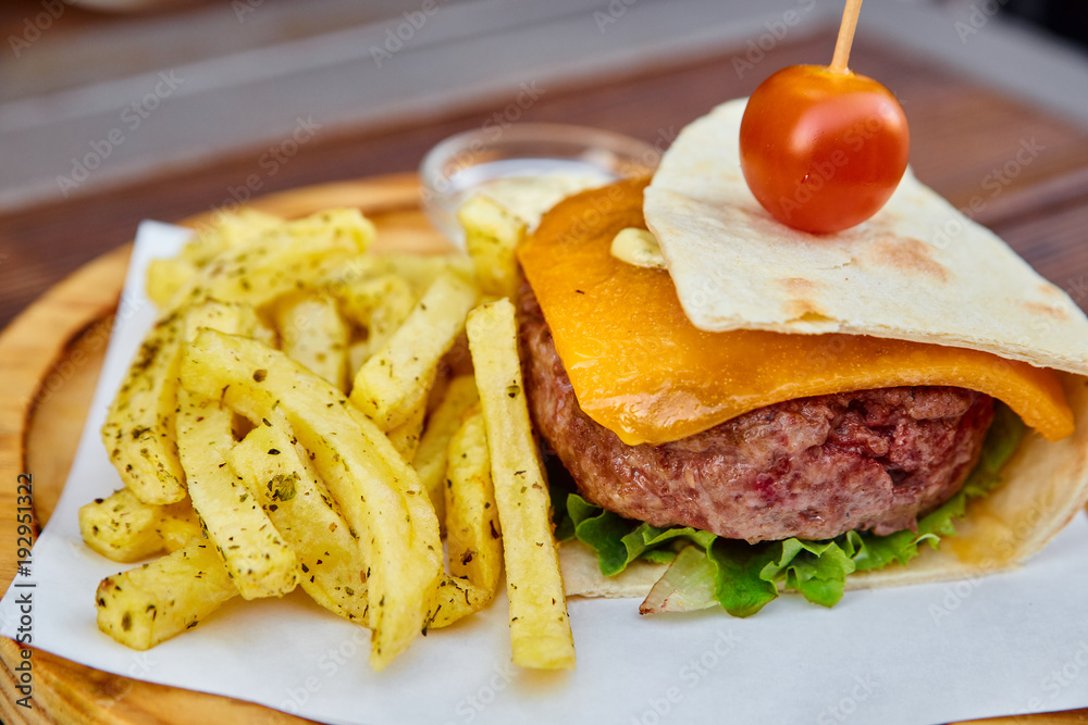 A huge juicy burger in a thin homemade pita bread with a sheet of green lettuce, melted cheddar cheese, veal cutlet and cherry tomato. Cheeseburger with French fries and white sauce on a wooden board.