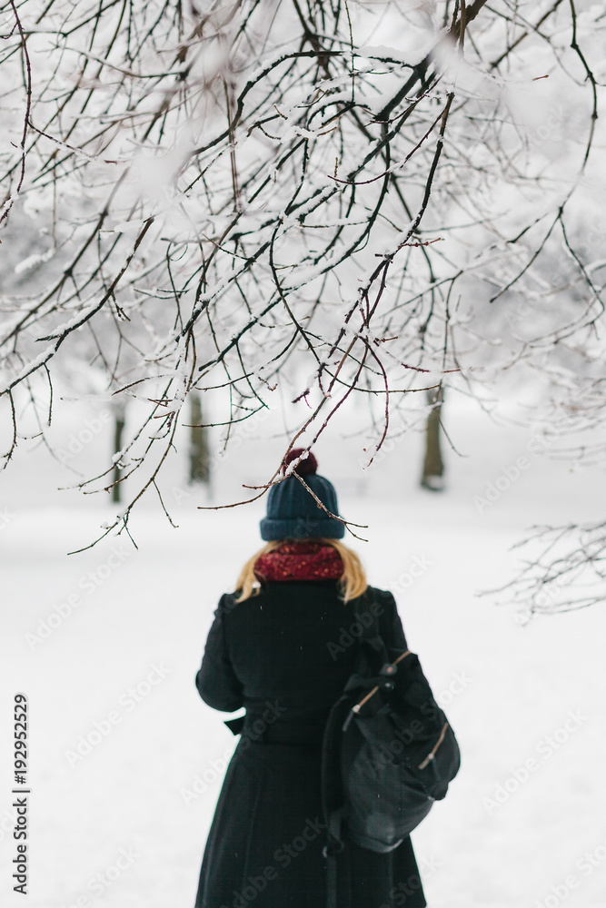 Young caucasian blonde girl in black coat and red knitted scarf walks in winter park Young caucasian blonde girl in black coat and red knitted scarf walks in winter park