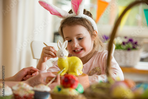 A mother and daughter celebrating Easter, painting bunny with brush in yellow color. Happy family smiling and laughing. Cute little girl in bunny ears preparing the holiday.